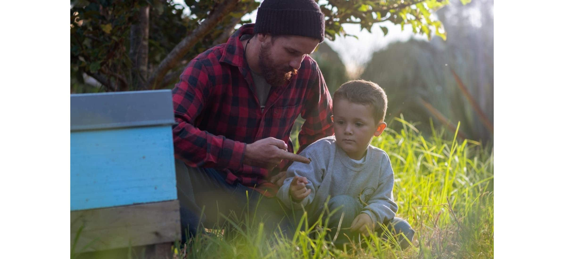 Rory sitting outside by a beehive, showing his young son a bee on his finger.